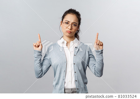 Skeptical, disappointed cute asian girl dont recommend this product, pointing fingers up with bothered, complaining and gloomy expression, have negative thoughts about company, grey background 81033524