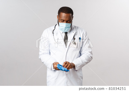Medicine, covid19, treatment and hospital concept. Serious-looking professional african-american doctor in face-mask get ready to treat patient sick, coronavirus symptoms, put on medical latex gloves Medicine, covid19, treatment and hospital concept. Serious-looking professional african-american doctor in face-mask get ready to treat patient sick, coronavirus symptoms, put on medical latex gloves 81034251