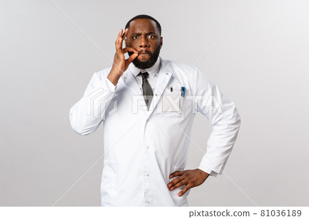Serious-looking angry and stressed african-american male doctor tired after working all night at hospital treating covid19 patients, making zip or lock lips gesture tell be quiet while he resting Serious-looking angry and stressed african-american male doctor tired after working all night at hospital treating covid19 patients, making zip or lock lips gesture tell be quiet while he resting 81036189