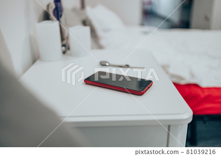 Black smartphone in a red case on the bedside table. Women's watch on the table in the bedroom against the background of the bed. Close up Black smartphone in a red case on the bedside table. Women's watch on the table in the bedroom against the background of the bed. Close up 81039216