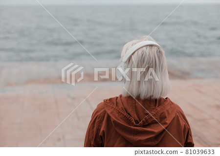 Back view of blonde woman wearing headphones listening music or podcast from smartphone application against the sea 81039633