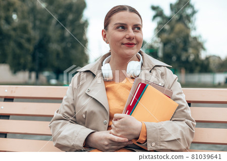 Young slender girl student with books and notebooks sits on a bench in a city park 81039641