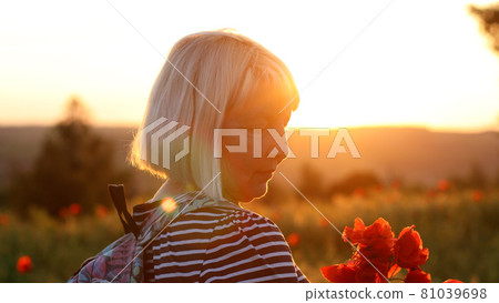 Adult Caucasian woman tourist with backpack and poppy flower bouquet at sunset in the field. Summer vacation. Happy girl with blond hair enjoying the sun. 81039698