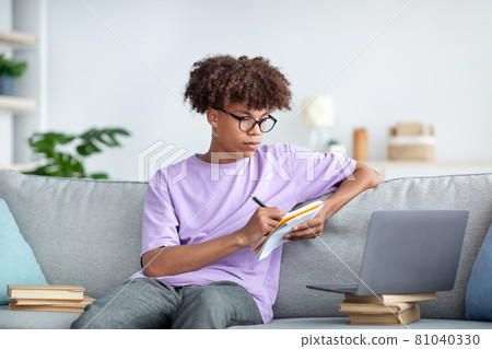 Focused Afro teen in glasses taking notes during online conference on laptop at home 81040330