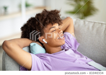 Peaceful black teen in earphones listening to music or audio book, lying on sofa at home 81040334