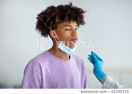 Doctor in rubber gloves making nasal coronavirus PCR test for African American teen, using sterile swab stick indoors 81040358