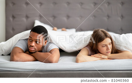 Sad Caucasian woman and her black husband lying apart on bed at home, looking in opposite directions Sad Caucasian woman and her black husband lying apart on bed at home, looking in opposite directions 81040360