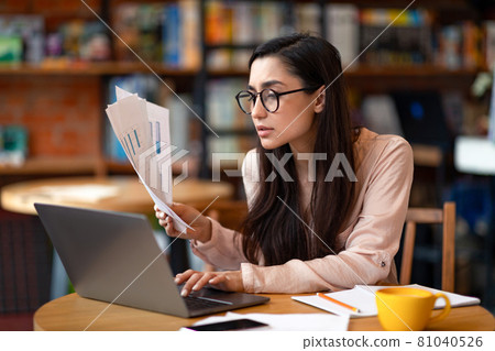 Successfull latin female entrepreneur working while sitting at cafe, using laptop and checking documents, copy space 81040526