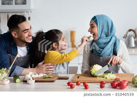 Cute little arab girl feeding muslim mom with cucumber slice in kitchen Cute little arab girl feeding muslim mom with cucumber slice in kitchen 81040539