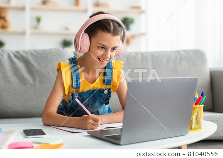 Smiling girl sitting at desk, using laptop, writing in textbook Smiling girl sitting at desk, using laptop, writing in textbook 81040556
