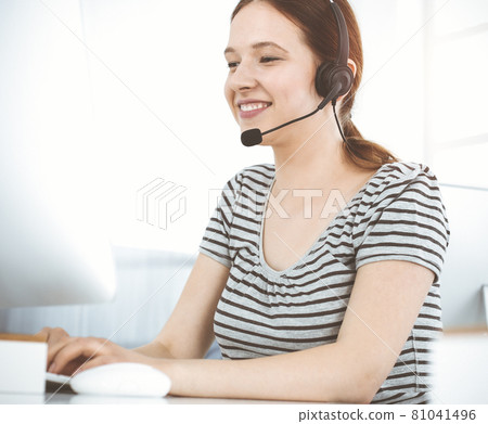 Casual dressed young woman using headset and computer while talking with customers online in sunny office. Call center Casual dressed young woman using headset and computer while talking with customers online in sunny office. Call center 81041496