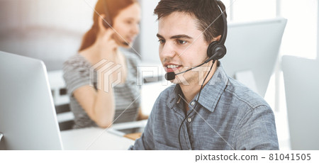 Casual dressed young man using headset and computer while talking with customers online in sunny office. Call center, business concept Casual dressed young man using headset and computer while talking with customers online in sunny office. Call center, business concept 81041505