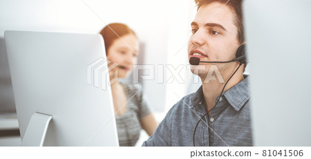 Casual dressed young man using headset and computer while talking with customers online in sunny office. Call center, business concept Casual dressed young man using headset and computer while talking with customers online in sunny office. Call center, business concept 81041506