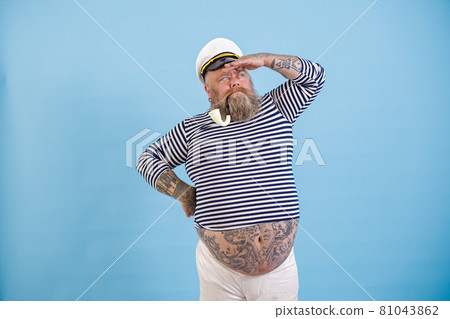 Plus size sailor with smoking pipe and hat looks into distance on light blue background Plus size sailor with smoking pipe and hat looks into distance on light blue background 81043862