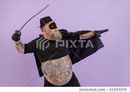 Fat man in Zorro costume with bare abdomen stands in bellicose pose on purple background 81043876