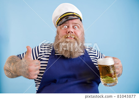 Joyful fat person in sailor suit holds beer and shows thumb up on light blue background Joyful fat person in sailor suit holds beer and shows thumb up on light blue background 81043877