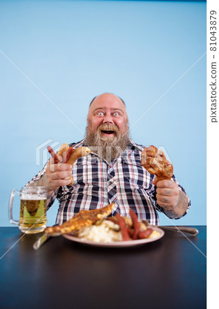 Excited obese man holds chicken leg and bunch of sausages at table in studio 81043879