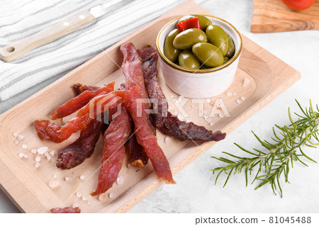 Jerky meat strips with spices, green olives, rosemary on wooden plate Jerky meat strips with spices, green olives, rosemary on wooden plate 81045488
