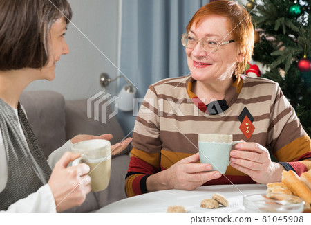 Two women drink tea and eat cakes near the New Year tree.  81045908