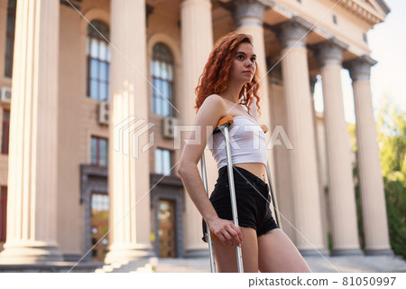 Young red-haired woman in shorts moves with crutches outdoors. 81050997
