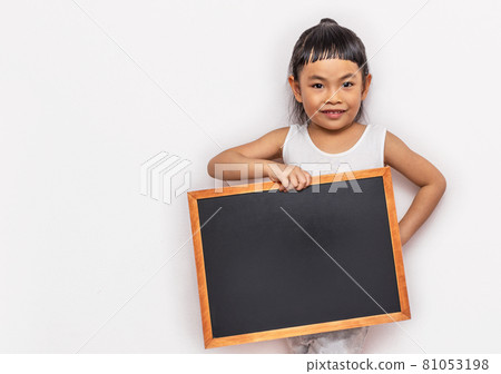Asian Little child girl holding a small blank black board with smiling face. Back to school concept. Wall background. Wearing white tank top. Looking at camera. 81053198