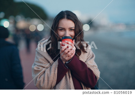 Young beautiful woman in coat and scarf with coffee cup outdoor. 81056019
