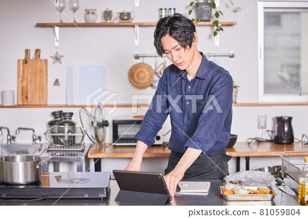 Image of a young man cooking in an apron who cooks while looking at the recipe on a tablet Image of a young man cooking in an apron who cooks while looking at the recipe on a tablet 81059804