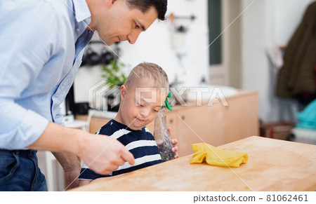 Father with happy down syndrome son indoors in kitchen, cleaning table. 81062461