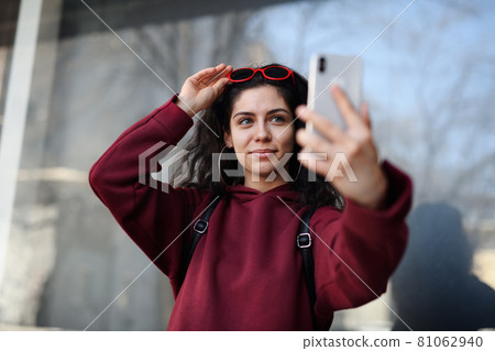 Portrait of young woman with smartphone standing outdoors on street in city, taking selfie. 81062940