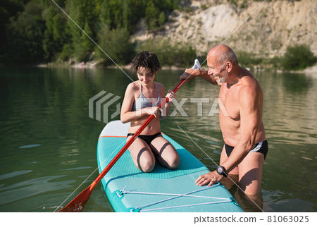 Happy preteen girl with grandfather on summer holiday by lake, paddleboarding. 81063025