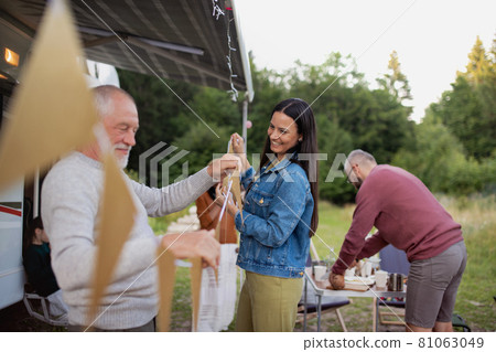 Multi-generation family preparing party by car outdoors in campsite, caravan holiday trip. 81063049