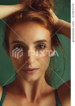 Close-up portrait of young beautiful red-headed woman without makeup isolated over dark green studio background. Natural beauty concept. Close-up portrait of young beautiful red-headed woman without makeup isolated over dark green studio background. Natural beauty concept. 81063570