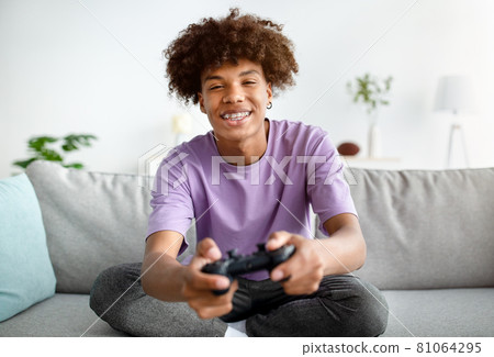Having fun at home. Cheerful black teen guy with joystick playing online computer games, sitting on couch indoors 81064295