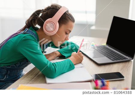 Girl in headset writing notes using laptop with empty screen Girl in headset writing notes using laptop with empty screen 81064388