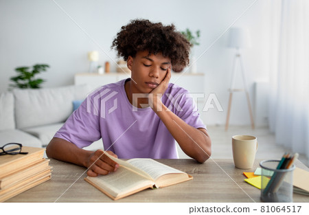 Bored black teenager sitting at desk, reading book, studying at home Bored black teenager sitting at desk, reading book, studying at home 81064517