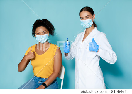 Young black woman with doctor in face masks approving of covid-19 vaccination, gesturing thumbs up before vaccine shot 81064663