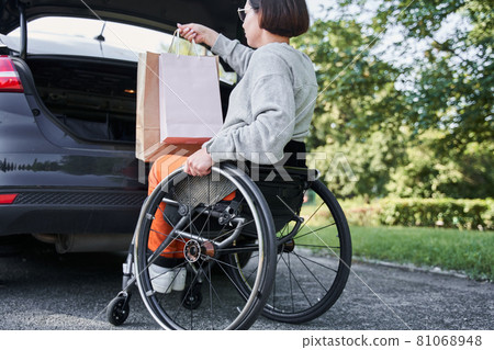 Girl sitting at the wheelchair is putting bags with purchases into car trunk 81068948