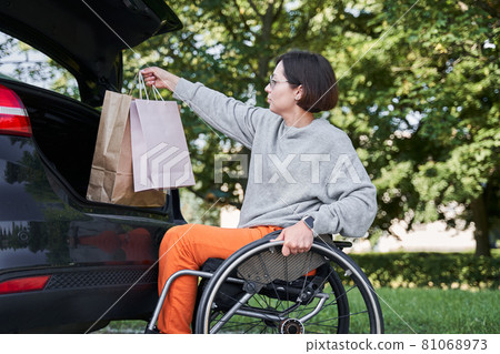 Woman sitting at her wheelchair and opening car trunk and putting shopping bags inside Woman sitting at her wheelchair and opening car trunk and putting shopping bags inside 81068973