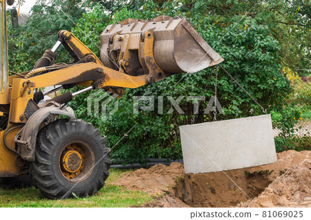 Hydraulic piston system of the excavator with a bucket, lowering into the pit on steel cables concrete sewer ring. Construction or repair of sewer home Hydraulic piston system of the excavator with a bucket, lowering into the pit on steel cables concrete sewer ring. Construction or repair of sewer home 81069025