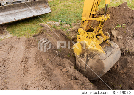 A bucket of excavator with a pile of earth digs a hole on the construction site. Industrial work A bucket of excavator with a pile of earth digs a hole on the construction site. Industrial work 81069030
