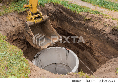 Excavator with a bucket, lowering into the pit on steel cables concrete sewer ring. Construction or repair of sewer home Excavator with a bucket, lowering into the pit on steel cables concrete sewer ring. Construction or repair of sewer home 81069035