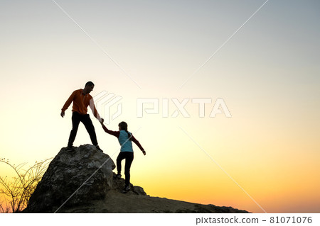 Man and woman hikers helping each other to climb a big stone at sunset in mountains. Couple climbing on a high rock in evening nature. Tourism, traveling and healthy lifestyle concept. Man and woman hikers helping each other to climb a big stone at sunset in mountains. Couple climbing on a high rock in evening nature. Tourism, traveling and healthy lifestyle concept. 81071076