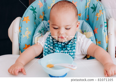 Serious baby sits at baby chair and looking to empty plate. Baby waiting for food in high chair Serious baby sits at baby chair and looking to empty plate. Baby waiting for food in high chair 81071105