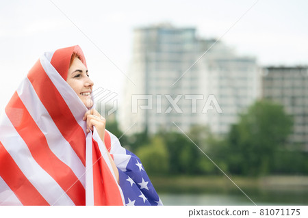 Portrait of young happy refugee woman with USA national flag on her head and shoulders. Positive muslim girl celebrating United States independence day. 81071175