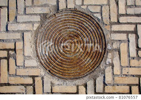 iron rusty round manhole on the sidewalk paved with stone tiles top view, old sewer hatch on a brick road. 81076557