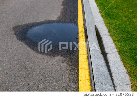 puddle on asphalt road with stormwater on the road side with a ditch for water after rain and a glade of green lawn along the highway. 81076558