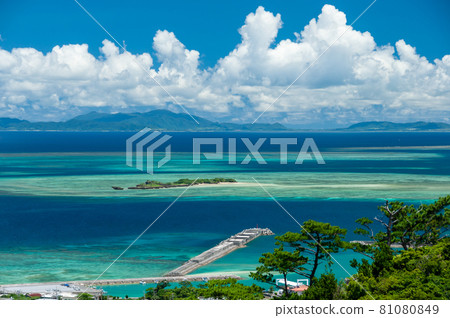 Stunning top view of a tropical island, pier extends over the blue and green gradient sea, blue sky, white clouds over Ishigaki island on horizon, green vegetation. Iriomote Island. Stunning top view of a tropical island, pier extends over the blue and green gradient sea, blue sky, white clouds over Ishigaki island on horizon, green vegetation. Iriomote Island. 81080849