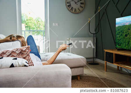 Woman watch TV set while luying on the sofa and switch channels with remote control 81087623