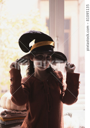 Halloween lady wearing witch cup. A woman in witch hat and Halloween pumpkin near the windows. Portrait of young female in orange sweater and traditional black witch cap 81095135