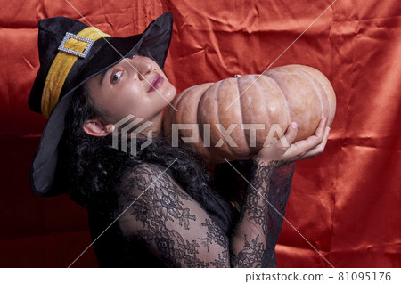Halloween lady wearing witch cup and holding a big Halloween pumpkin. A woman in witch hat and Halloween pumpkin over orange background. Portrait of young female in traditional black witch cap 81095176